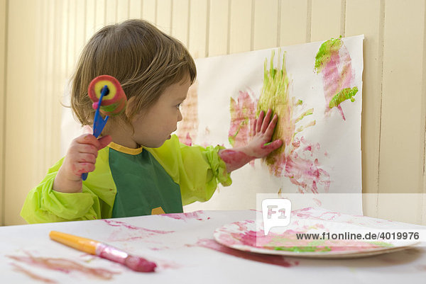 Mädchen  2 Jahre alt mit Malschürze aus Plastik  malt in einem Spielzimmer mit Pinsel und Malrolle  Walze