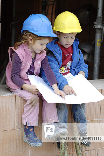 Zwei Kinder  Geschwister  mit Bauhelmen und Bauplan in einem Rohbau auf einer Baustelle