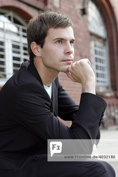 Portrait eines jungen Mannes im Anzug  der vor einem Industriegebäude aus roten Backsteinen sitzt und nachdenkt