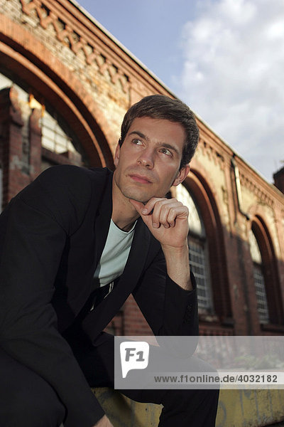 Portrait of a young man wearing a suit  sitting in front of an industrial building made of red bricks looking into the distance  thinking