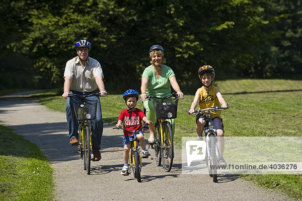 Family with 2 children riding bicycles through a park  wearing helmets