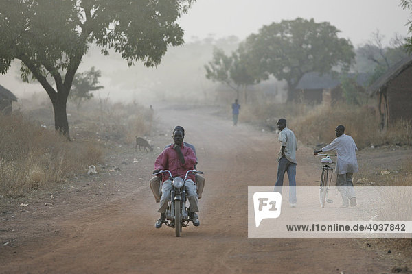 Männer mit Fahrrad und Motorrad auf der Dorfstraße  im morgendlichen Dunst  Houssere Faourou  Kamerun  Afrika