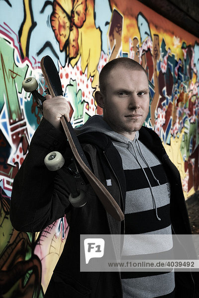 Junger Mann mit Skateboard vor einer Graffitiwand  Portrait