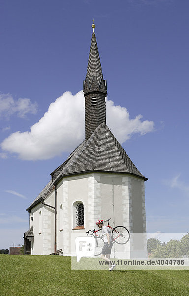 Radfahrer trägt Rennrad vor Kirche  Kapelle in Kleinhartpenning in der Nähe von Holzkirchen  Oberland  Bayern  Deutschland  Europa
