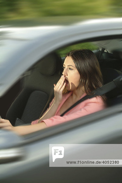 Junge Frau sitzt müde und gähnend am Steuer ihres Autos