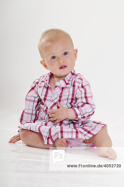 Baby  9 Monate  mit rosa kariertem Kleid sitzt auf dem Boden  aufmerksam