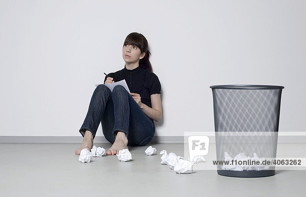 Junge dunkelhaarige Frau sitzt an eine Wand gelehnt zwischen geknülltem Papier auf dem Boden und überlegt
