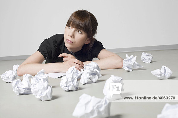 Young  dark-haired woman lying amongst screwed-up balls of paper  deliberating with a pen in her hand