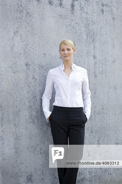 Junge blonde Frau im Business-Look steht vor einer grauen Wand