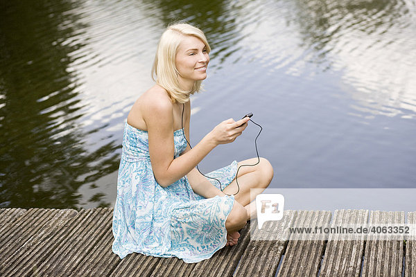 Junge blonde Frau sitzt in einem Kleid auf einem Steg am Wasser und hört Musik