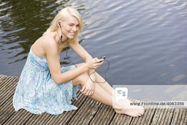 Junge blonde Frau sitzt in einem Kleid auf einem Steg am Wasser und hört Musik