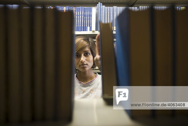 Junge dunkelhaarige Frau  Studentin  greift nach einem Buch aus einem Bücherregal in der Bibliothek
