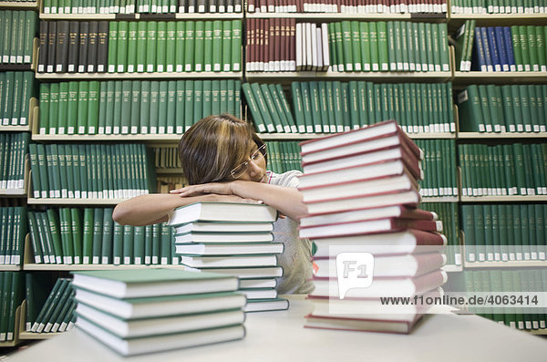 Junge dunkelhaarige Frau  Studentin  sitzt schlafend mit vielen Büchern an einem Tisch vor einem Bücherregal in einer Bilbiothek