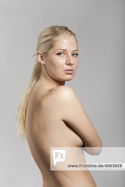 Junge blonde Frau steht mit unbekleidetem Oberkörper vor Grau und verdeckt mit ihren Armen die Brust