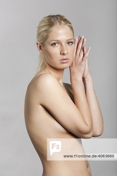 Young blonde woman standing topless in front of grey  covering her breasts with her arms