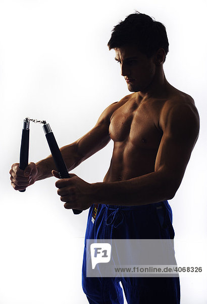 Young fighter with bare upper body holding nunchaku  backlight