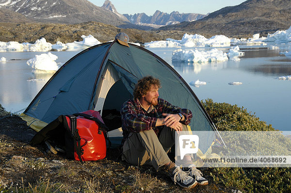 Junger Mann  Abenteurer sitzt vorm Zelt  hinten Eisberge  Paarnakajiit  Sermilik Fjord  Ostgrönland  Grönland  Arktis
