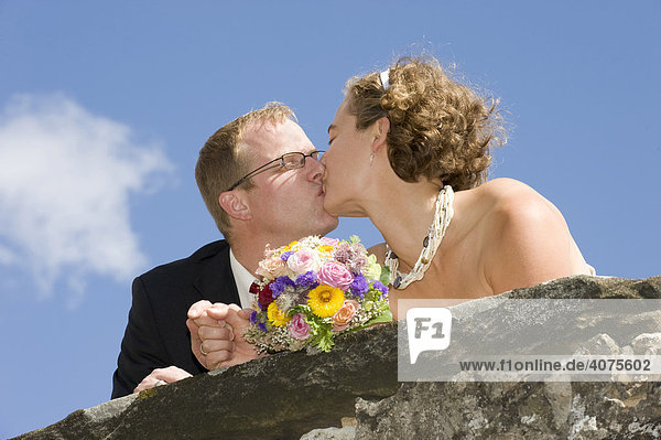 Junges Hochzeitspaar mit einem Brautstrauß küsst sich auf Burgmauer