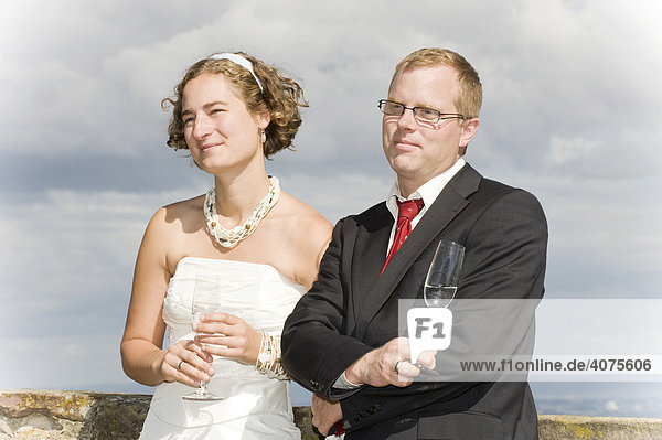 Young bride and groom holding champagne glasses lean against a castle wall