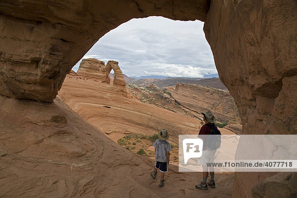 John West und sein Sohn Joey  9  unter einem kleinen Felsenbogen mit Blick auf den Delicate Arch Felsbogen  Arches Nationalpark  Moab  Utah  USA
