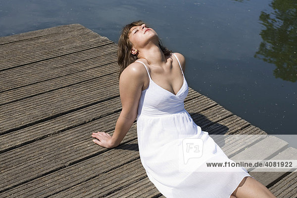 Frau im weißem Kleid sitzt auf einem Holzsteg und genießt die Sonne
