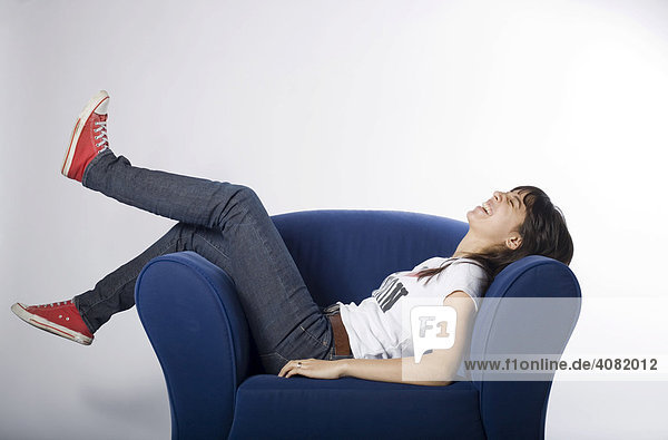Junge Frau liegt bequem auf einem blauen Sessel und lacht
