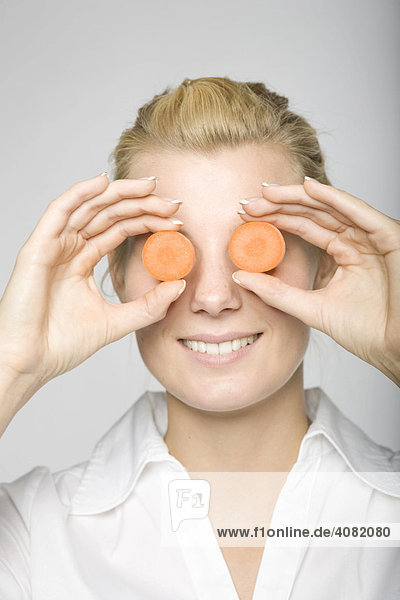 Junge blonde Frau hält sich zwei Karottenscheiben vor die Augen