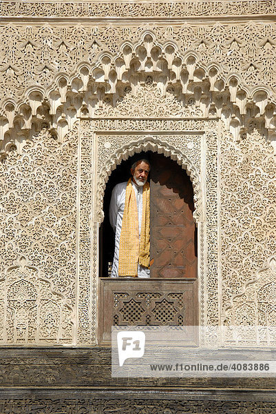 Man wearing traditional cloth looks out of a window decorated with rich ornaments Medersa Bou Inania Fes El-Bali Morocco