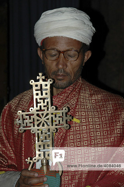 Priester mit rotem Umhang und weißer Kopfbedeckung trägt kunstvoll gearbeitetes Lalibela Kreuz Felsenkirchen Lalibela Äthiopien