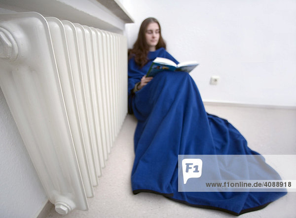 Junge Frau sitzt in Decke gehüllt an der Heizung  um Energiekosten zu sparen.