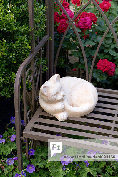 Gartendekoration  Steinkatze liegt auf einer Metallbank im Garten  vor Buchs buxus  Rosen rosa und Storchschnabel geranium