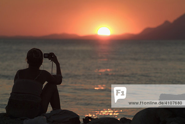 Woman taking a photograph of a sunset over the ocean with a digital camera