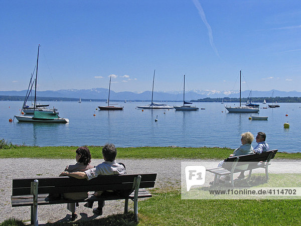 BRD Germany Bavaria Upper Bavaria Tutzing at the Starnberger Lake Holiday Region Recreation Area for Munich Upper Bavarian Watering Lake Visitors are sitting on Benches at the Beachside View to the Lake and Boats and Mountain Range Calm Frame Holiday Frame Elder People enjoy Calmness and View