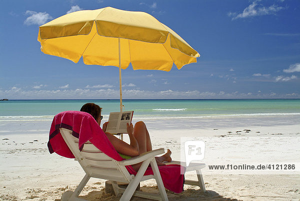 Junge Frau liest ein Buch im Liegestuhl unter gelbem Sonnenschirm  Insel Praslin  Seychellen  Afrika