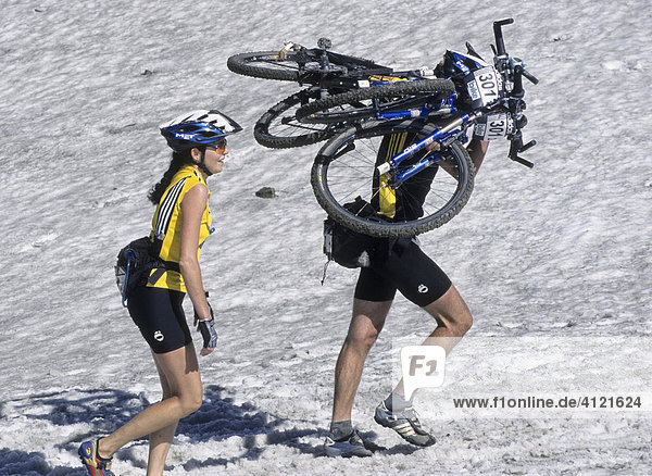Zwei Teilnehmer der Adidas Bike Transalp Challenge Mountainbikerennen Mann trägt zwei Räder auf den Schultern Frau mit gelbem Trikot und Helm geht lächelnd daneben her Südtirol Italien Europa