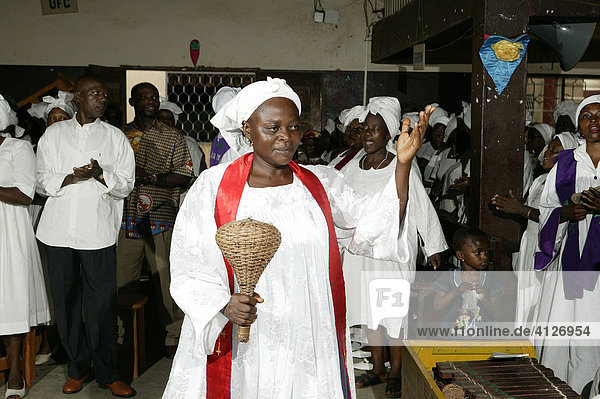 Woman dressed in white robes and red sash making music with a wicker rattle during a church service in Douala  Cameroon  Africa