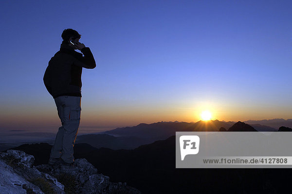 Mann mit Handy steht bei Sonnenaufgang auf einem Berggipfel  Breitenstein  Bayerische Voralpen  Wendelsteingruppe  Oberbayern  Bayern  Deutschland