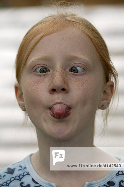 7-jähriges Mädchen mit roten Haaren macht Grimassen  streckt Zunge raus und schielt