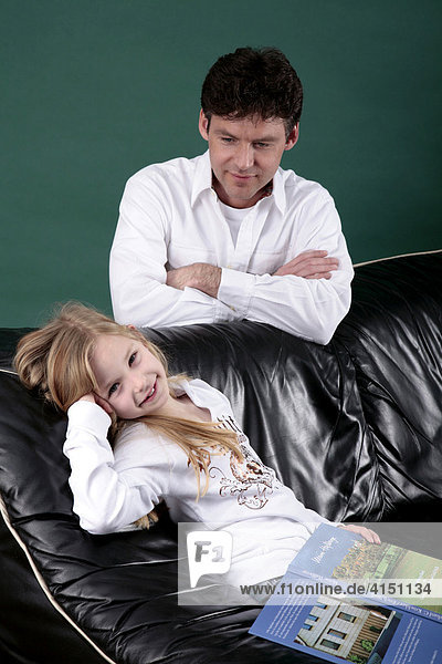 Tochter sitzt mit Buch auf dem Sofa während der Vater dahinter auf die Tochter blickt