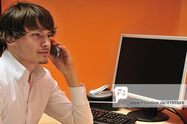 Junger Mann sitzt am Schreibtisch mit Computer und telefoniert