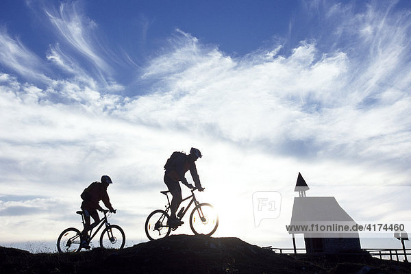 Silhouette von zwei Mountainbikern in den Bergen vor einer kleinen Kapelle