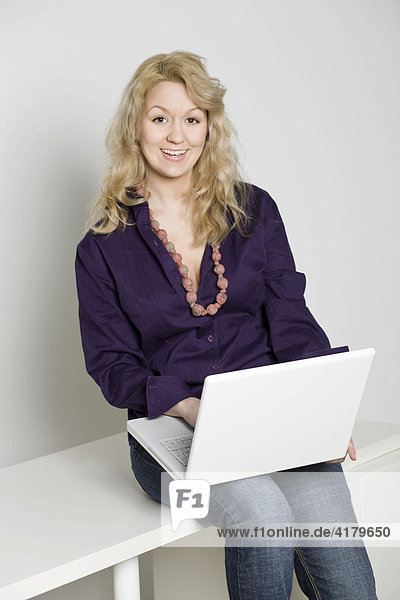 Junge bonde Frau sitzt auf einem Bürotisch und arbeitet mit einem weißen Notebook
