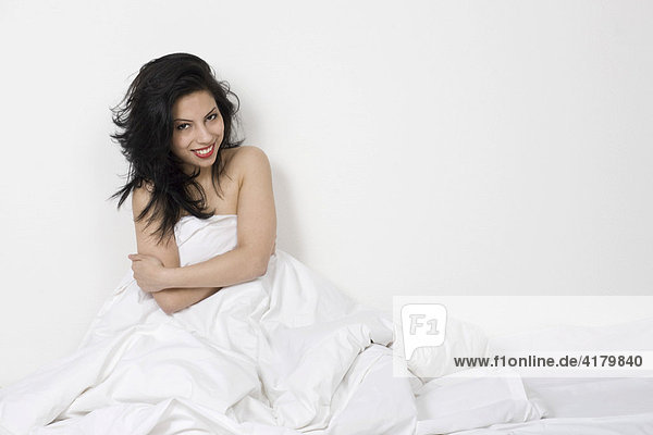 Junge dunkelhaarige Frau sitzt lächelnd mit einer weißen Bettdecke um den Körper gewickelt im Bett