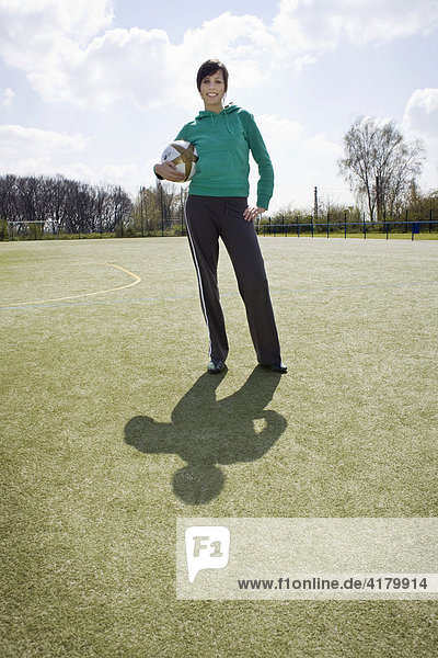 Junge Frau steht auf einem Sportplatz und hält einen Fußball fest
