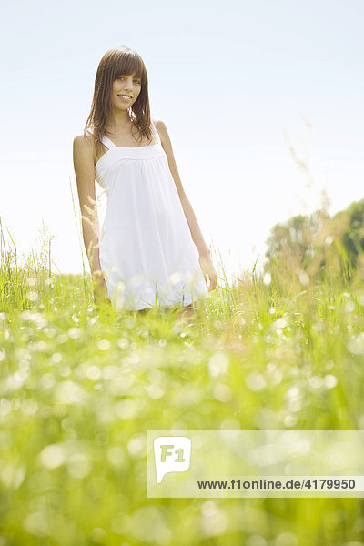 Junge dunkelhaarige Frau in einem weißen Kleid steht auf einer sommerlichen Wiese und blickt freundlich in die Kamera