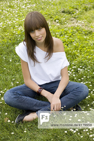 Young  dark-haired woman wearing jeans and a white top  sitting in a sommerly meadow  looking friendly at camera