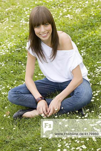 Junge dunkelhaarige Frau mit Jeans und weißem Top sitzt auf einer sommerlichen Wiese und blickt freundlich in die Kamera
