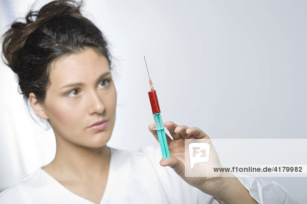 Junge Frau mit weißem Laborkittel hält eine Spritze mit roter Flüssigkeit in den Händen und blickt konzentriert