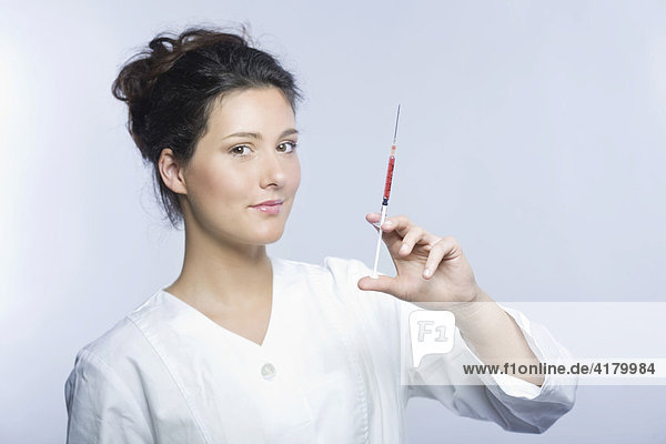 Junge Frau mit weißem Laborkittel hält eine Spritze mit roter Flüssigkeit in den Händen