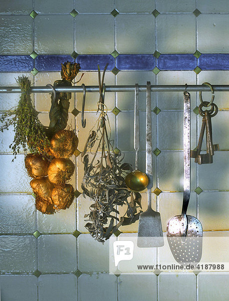 Kachelwand mit Knoblauchzopf  getrocknetem Salbei und Küchengeräten sowie altem Schlüssel